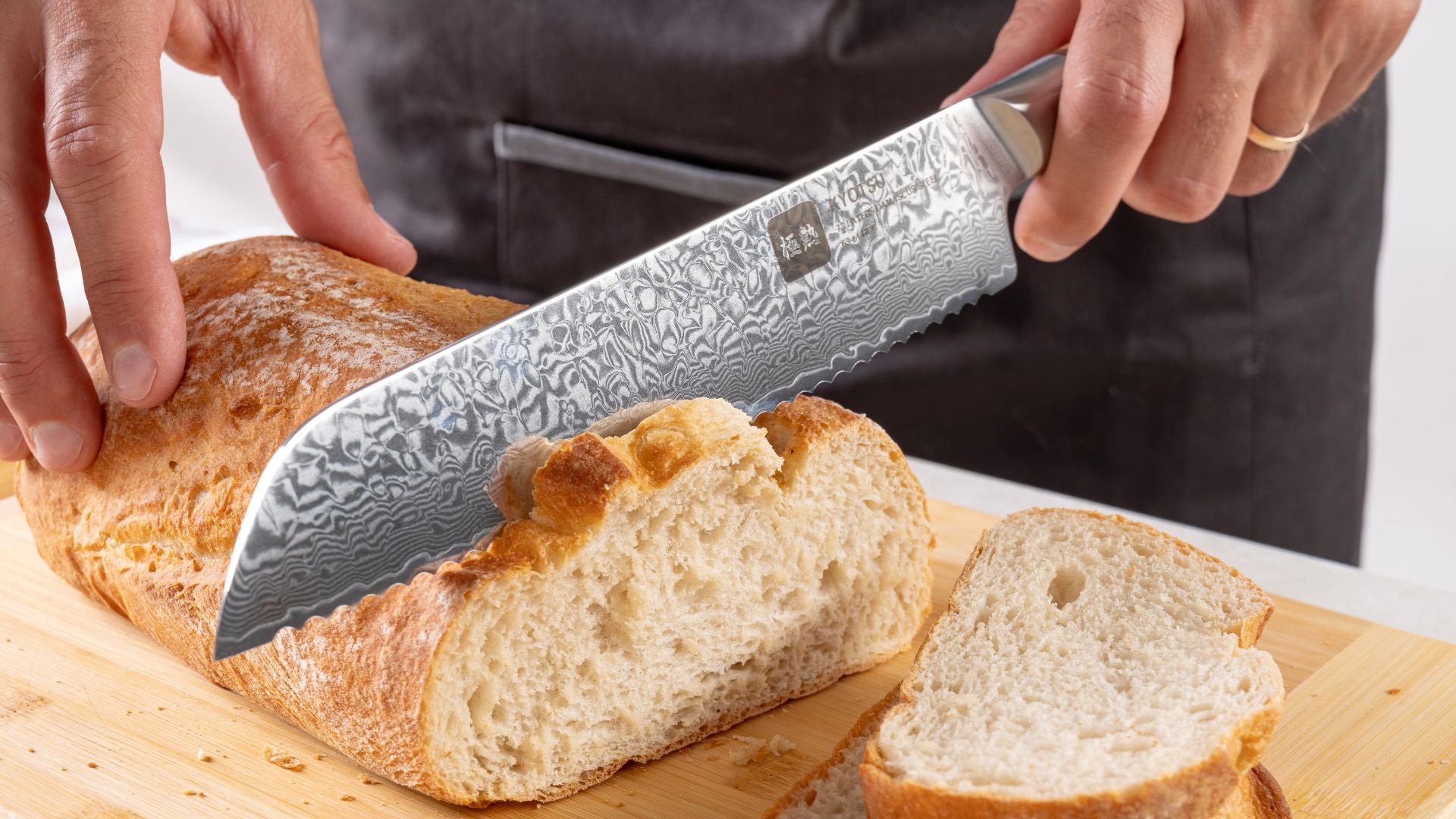 Eine Person schneidet mit dem Kyotsu Brotmesser aus Damaststahl ein Brot auf einem Holz-Schneidebrett – hochwertiges Messer in Aktion.
