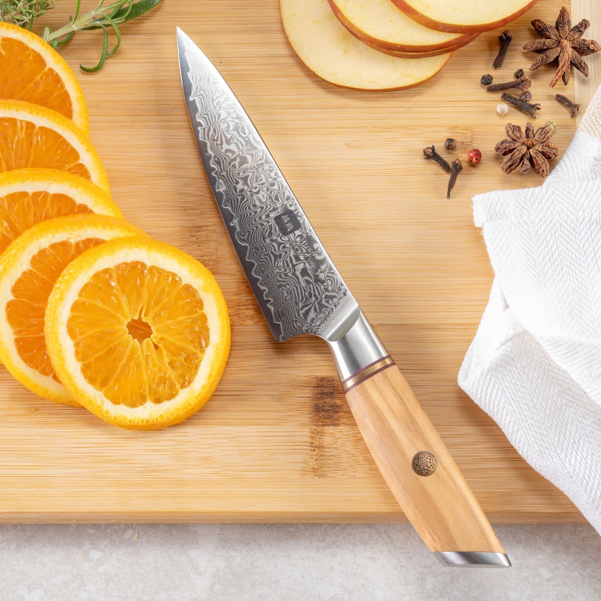Kyotsu Allzweckmesser aus Damaststahl auf Holz-Schneidebrett mit frischen geschnittenen Orangen, Äpfel und Nelken – stilvolle Produktpräsentation für hochwertige Küchenmesser.