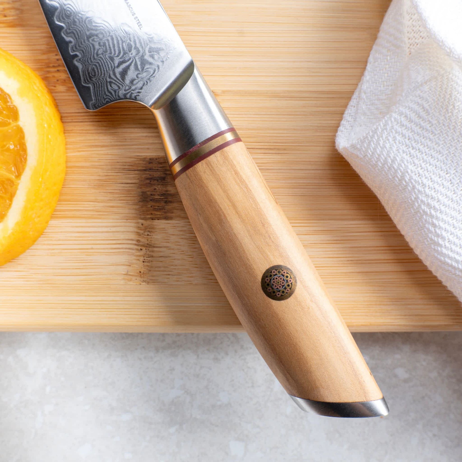 Nahaufnahme des Olivenholzgriffes vom Kyotsu Allzweckmesser aus Damaststahl – daneben frisch geschnittener Orangenschnitz.