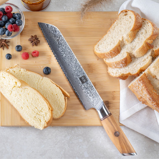 Kyotsu Brotmesser aus Damaststahl auf Holz-Schneidebrett mit frischem Brot und Früchte – stilvolle Produktpräsentation für hochwertige Küchenmesser.