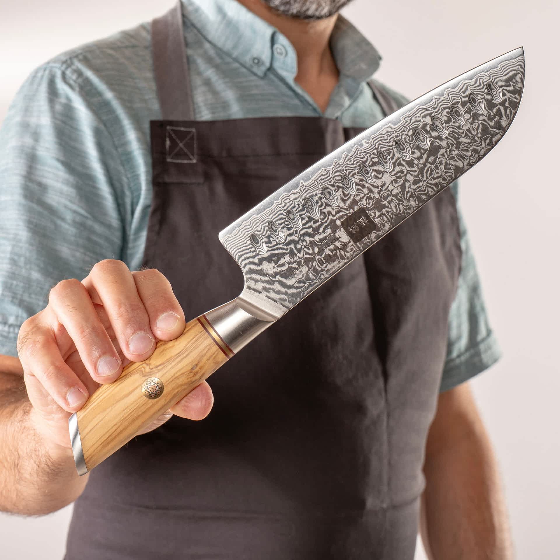 Person mit Schürze präsentiert Kyotsu Santoku-Messer aus Damaststahl.