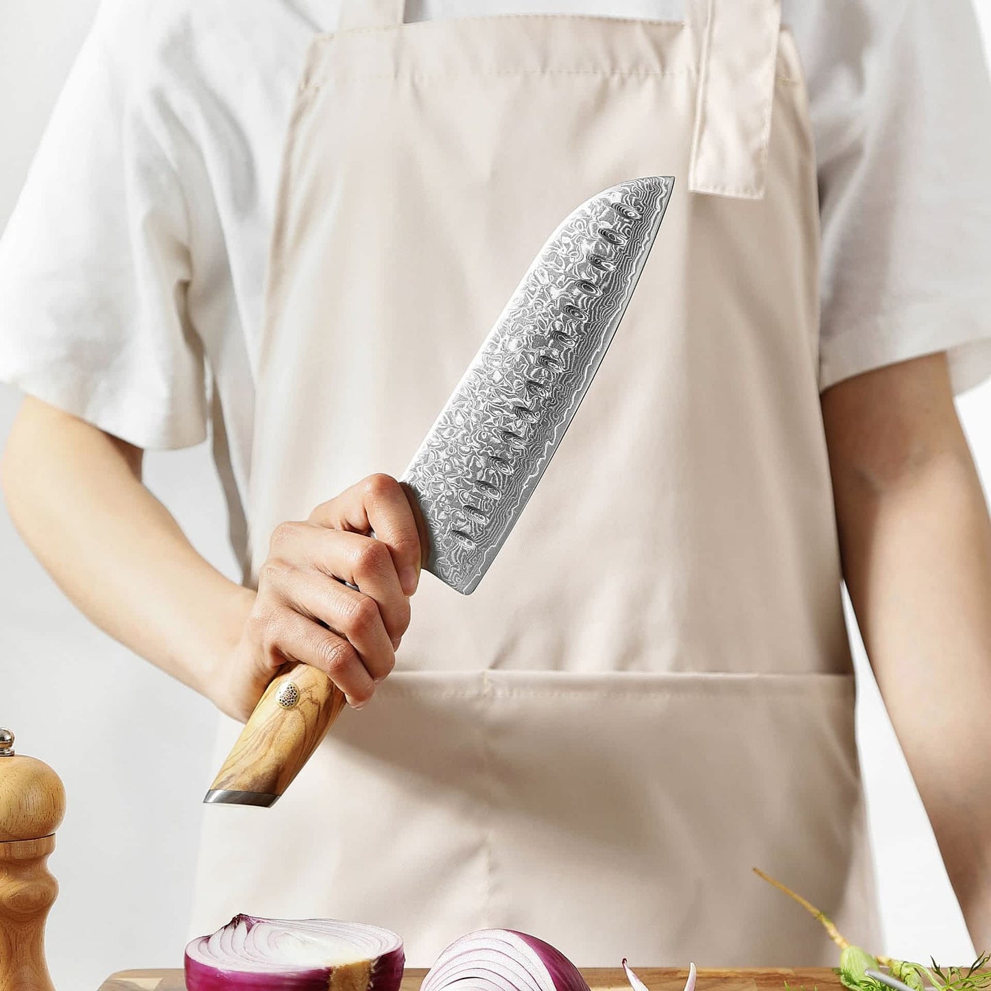 Person mit Schürze präsentiert Kyotsu Santoku-Messer aus Damaststahl – Holzbrett mit frischem Gemüse im Vordergrund.