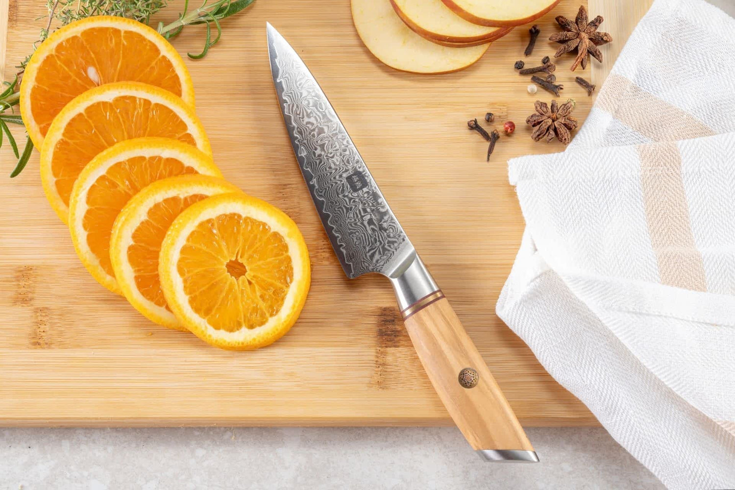 Kyotsu Allzweckmesser aus Damaststahl auf Holz-Schneidebrett mit frischen geschnittenen Orangen, Äpfel und Nelken – stilvolle Produktpräsentation für hochwertige Küchenmesser.