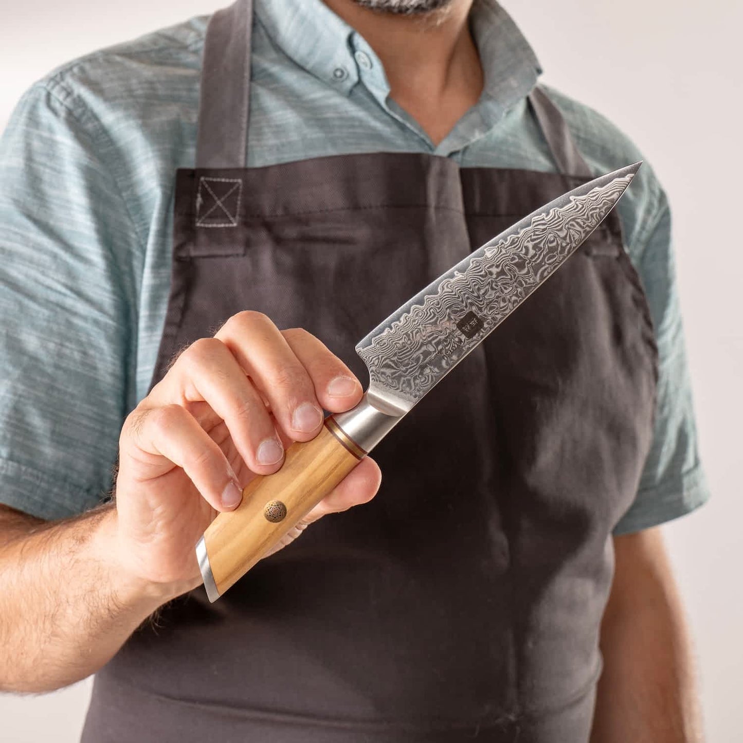 Ein Mann mit Kochschürze hält das japanische Allzweckmesser aus Damaststahl in die Kamera. Auf der Klinge ist die Gravur von Kyotsu inkl. Logo ersichtlich.