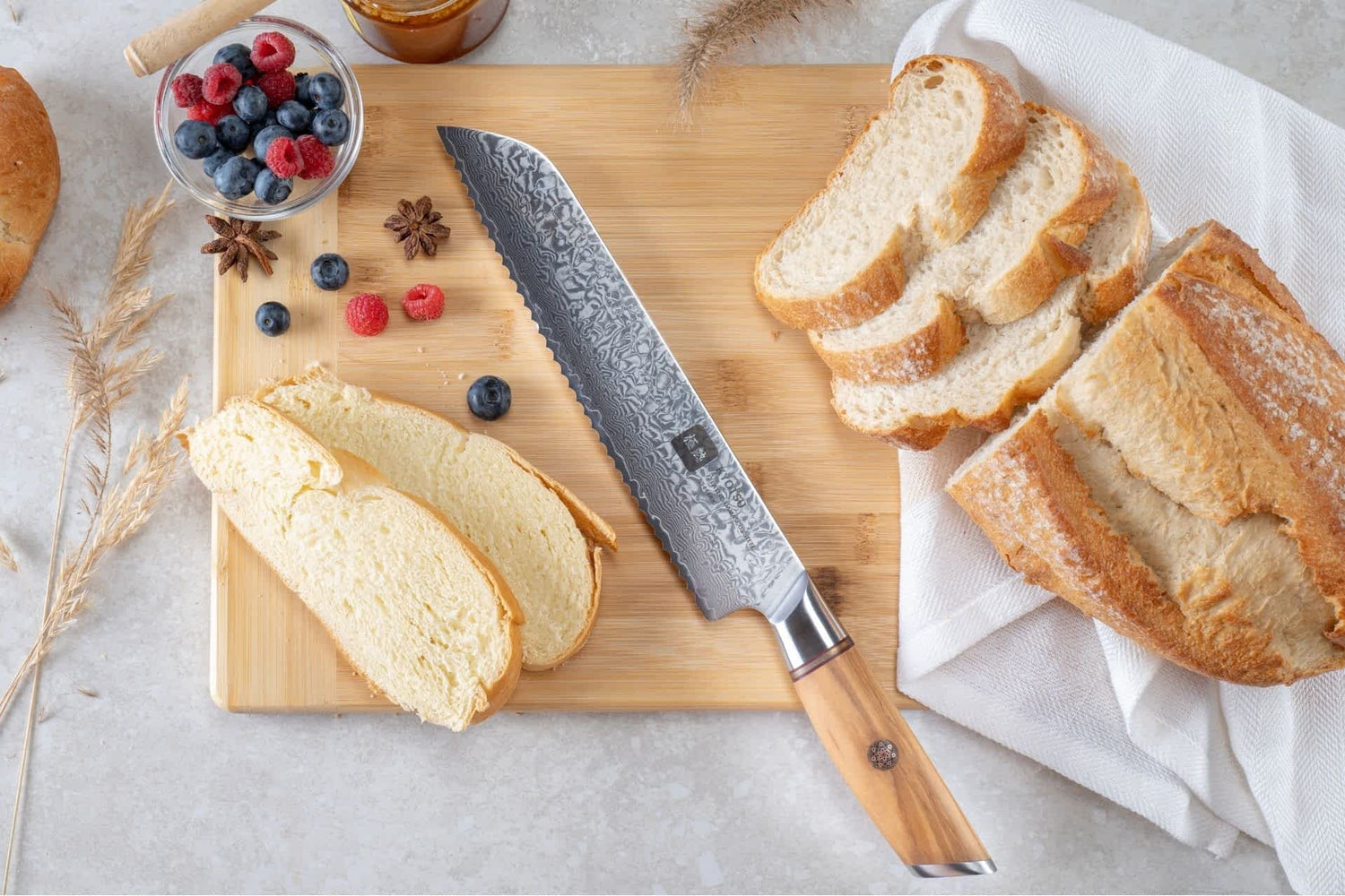 Kyotsu Brotmesser aus Damaststahl auf Holz-Schneidebrett mit frischem Brot und Früchte – stilvolle Produktpräsentation für hochwertige Küchenmesser.