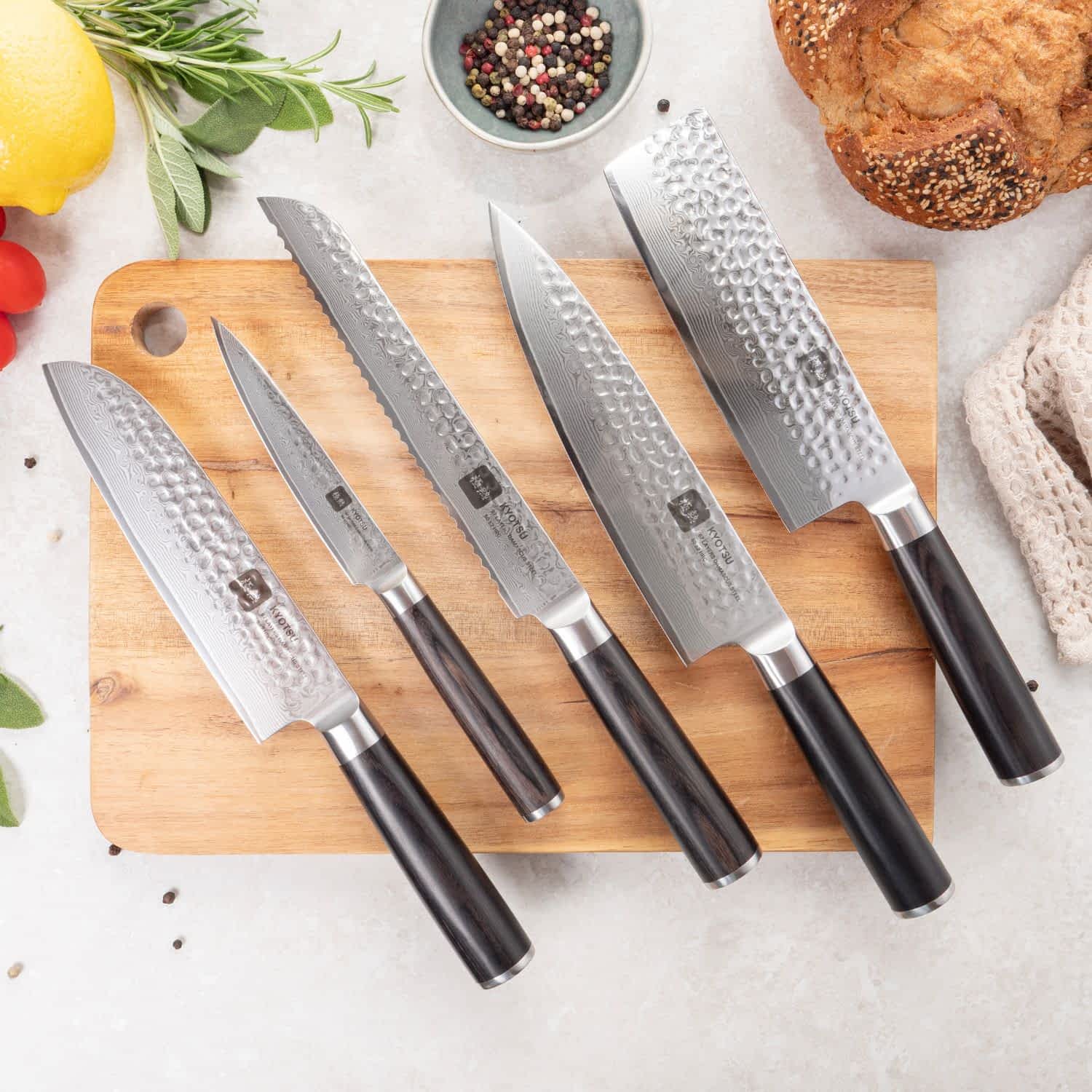 Auf einem Holzbrett liegen fünf Damastmesser. Daneben sind Produkte wie Tomaten, Zitrone, Pfefferkörner, Kräuter und Brot als Dekoration platziert.