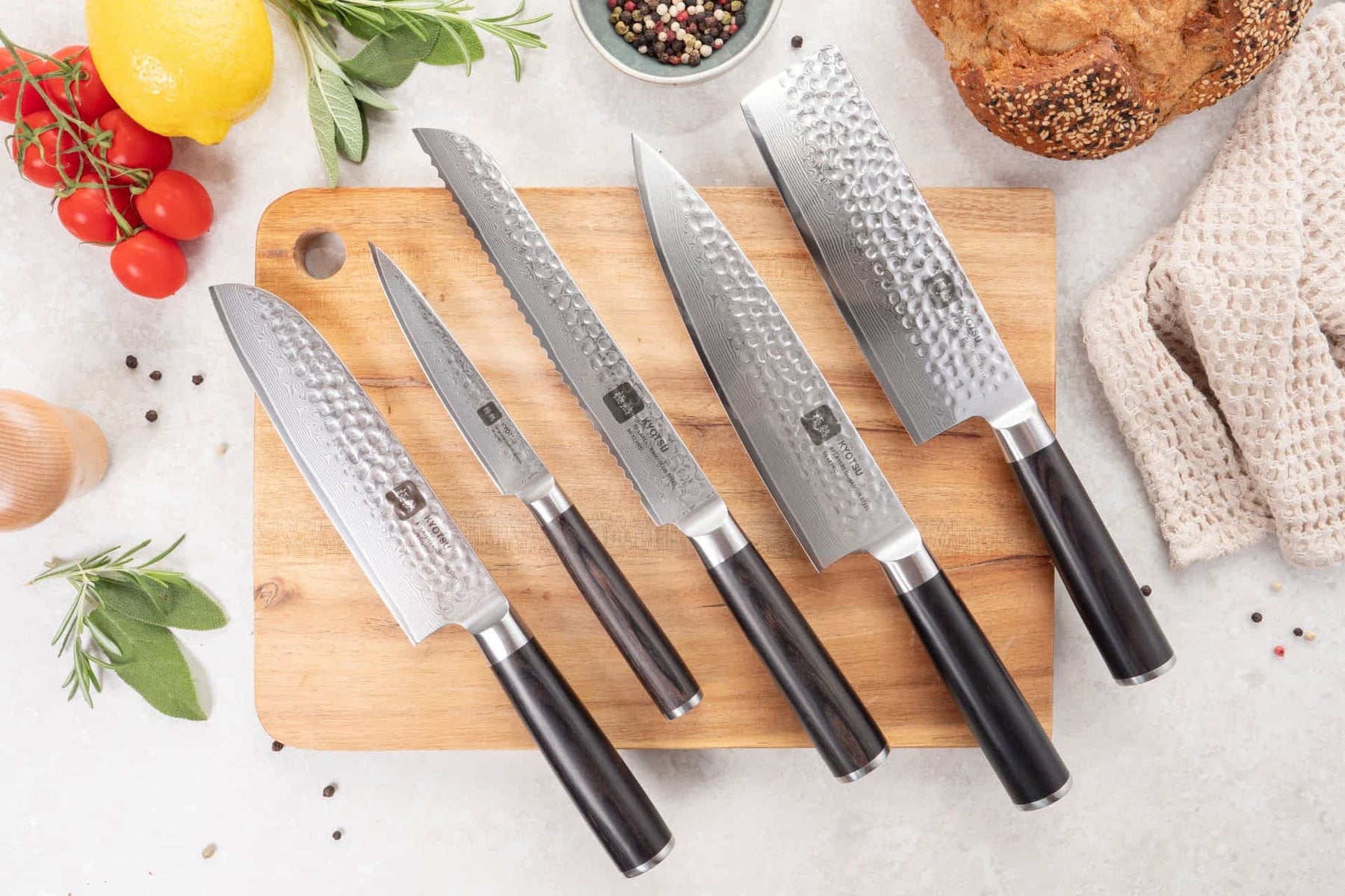 Auf einem Holzbrett liegen fünf Damastmesser. Daneben sind Produkte wie Tomaten, Zitrone, Pfefferkörner, Kräuter und Brot als Dekoration platziert.