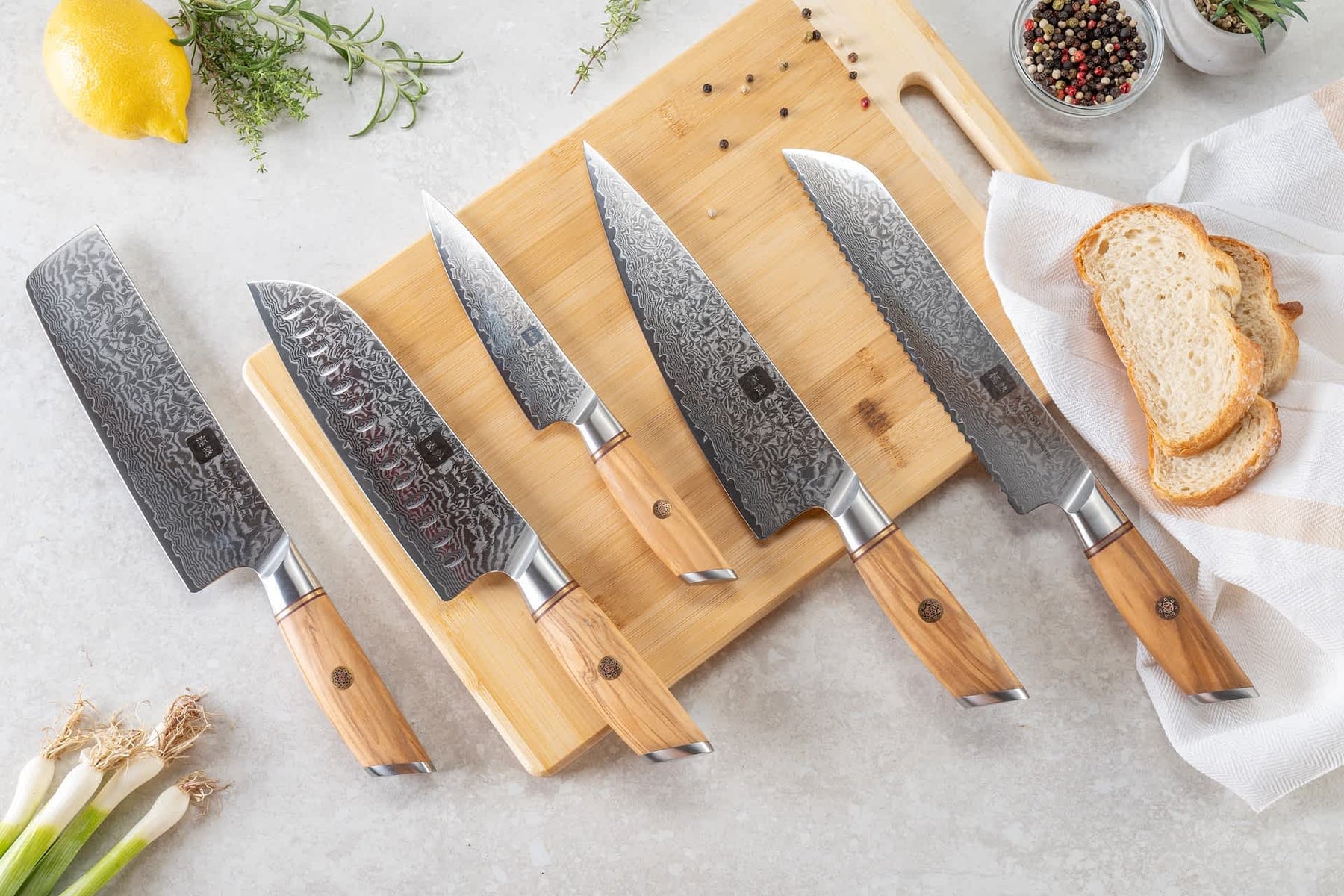 Auf einem Holzbrett liegen fünf Damastmesser. Daneben sind Produkte wie Tomaten, Zitrone, Frühlingszwiebeln, Knoblauch, Pfefferkörner, Kräuter und Brot als Dekoration platziert.