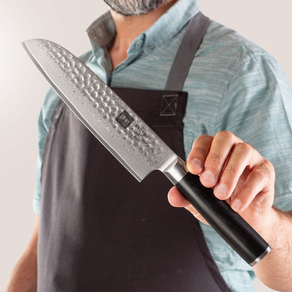 Person mit Schürze präsentiert Kyotsu Santoku-Messer aus Damaststahl.