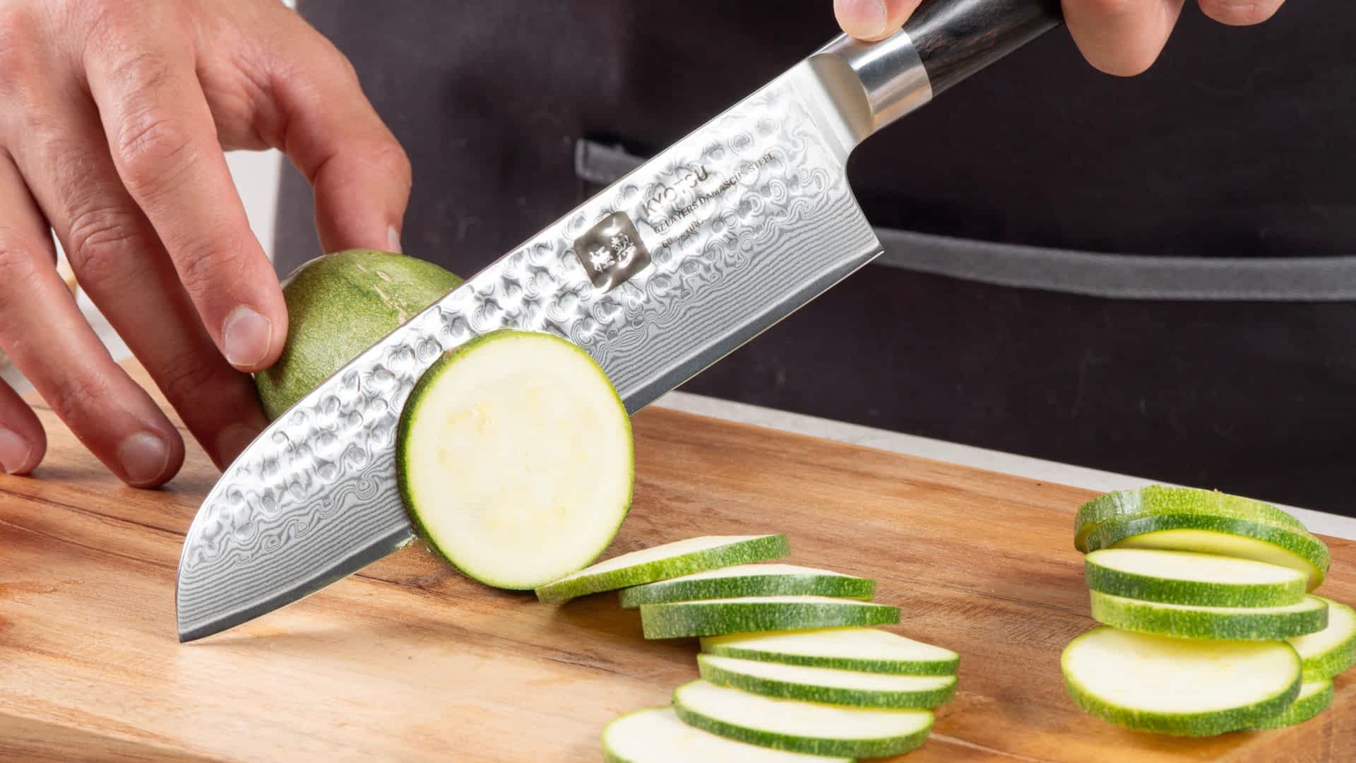 Eine Person schneidet mit dem Kyotsu Santoku-Messer aus Damaststahl Zucchini auf einem Holz-Schneidebrett – hochwertiges Messer in Aktion.